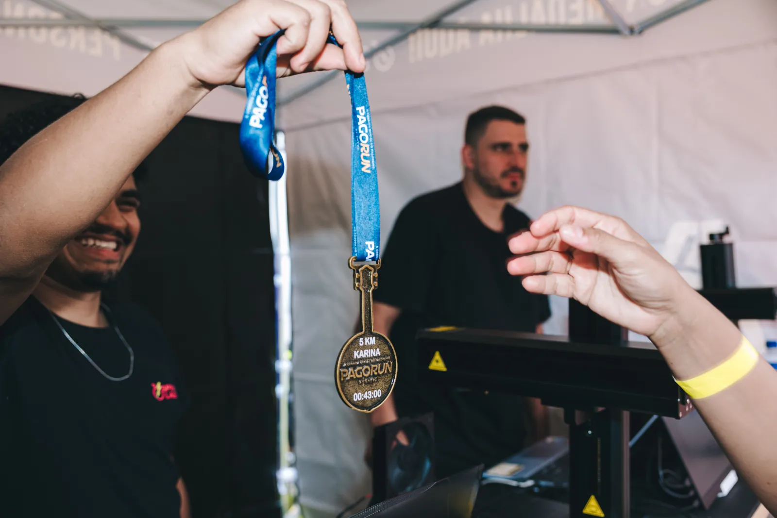 Pessoa sorri e ergue uma medalha da Pagorun gravada com 5 km, o nome Karina e o tempo 00:43:00, enquanto outra mão se estende para recebê-la na tenda de gravação