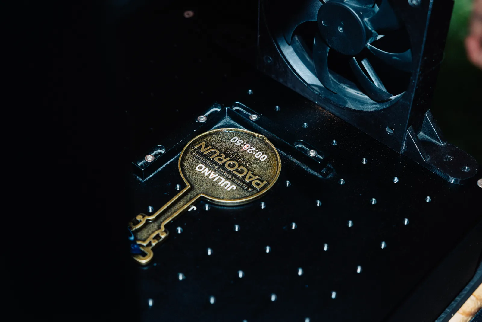 Medalha dourada da Pagorun gravada com o nome Juliano e o tempo de prova, fixada sobre a mesa preta da máquina de gravação a laser