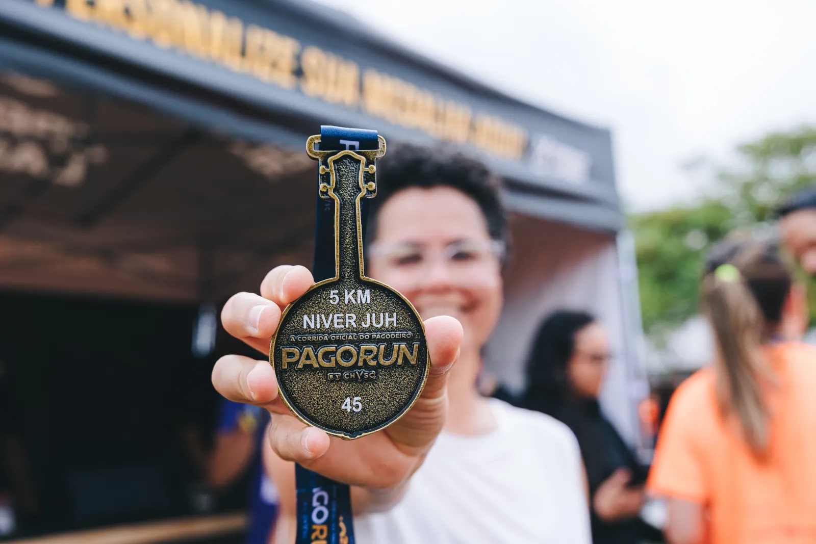 Pessoa sorrindo mostra para a câmera uma medalha da Pagorun gravada com 5 km e Niver Juh, em frente à tenda de personalização de medalhas